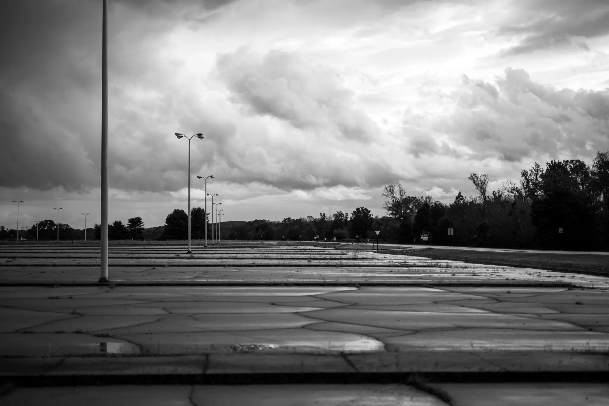 An empty parking lot with tall light poles under heavy storm cloud, trees along the far edge