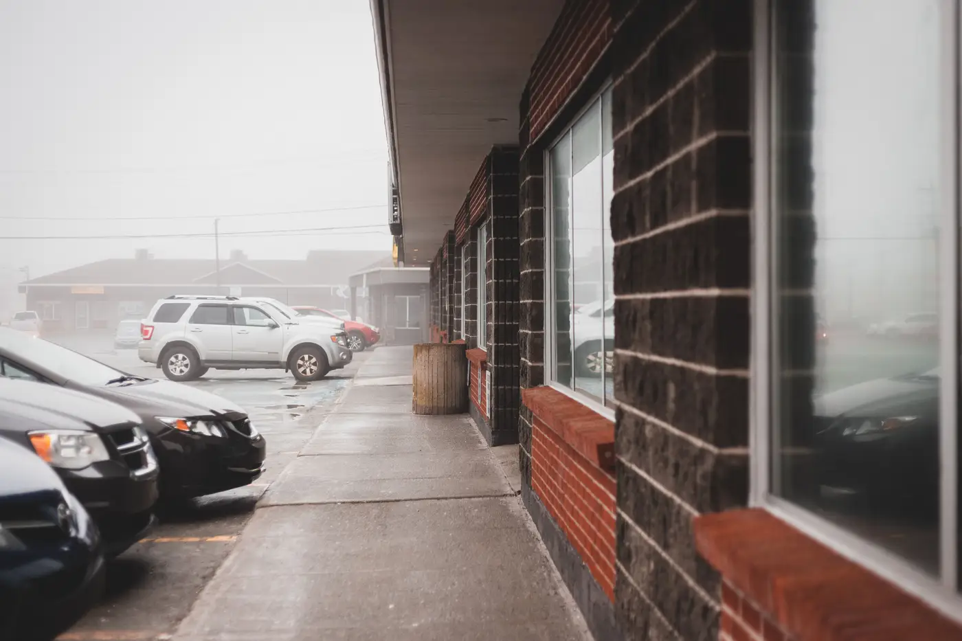 Cars parked along a covered walkway outside a brick commercial storefront on a rainy day.